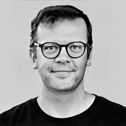 Black and white portrait of a man wearing glasses, smiling, with short dark hair, dressed in a black shirt.