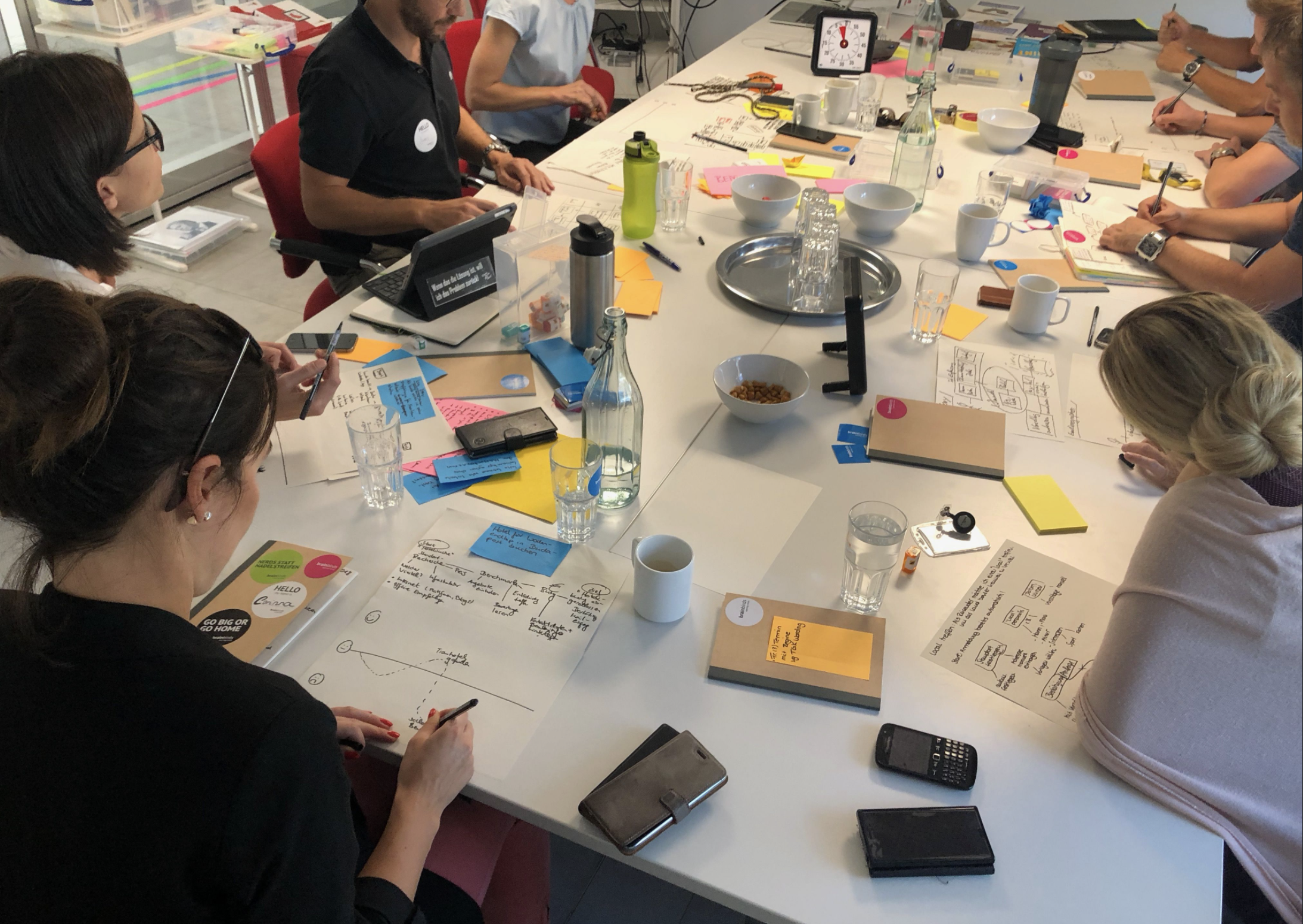 A group of people sitting around a large conference table engaged in a meeting or workshop. The table is filled with notebooks, papers, pens, sticky notes, glasses of water, and electronic devices such as smartphones and tablets. There are also bowls