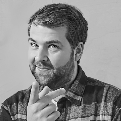 Black and white photo of a man with a beard and short hair, wearing a checkered shirt, smiling with his hand near his chin.