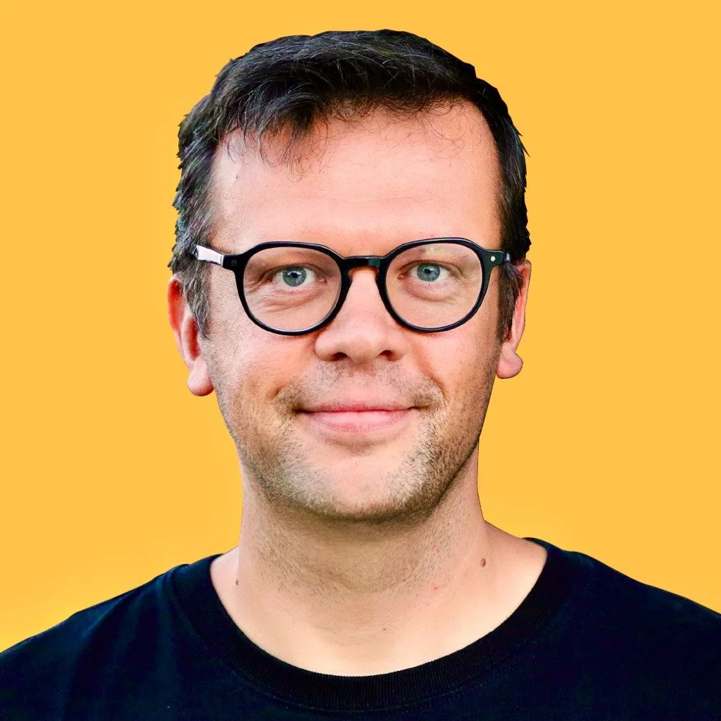 A man with glasses smiling against a yellow background.