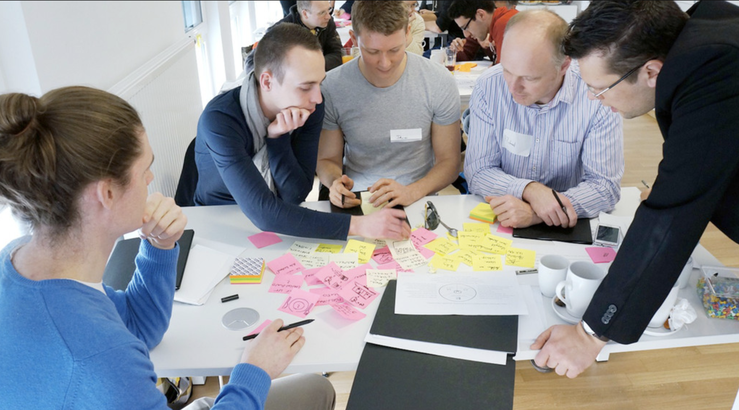 Group of five people collaborating at a table, reviewing notes and sticky notes, in a bright office environment.