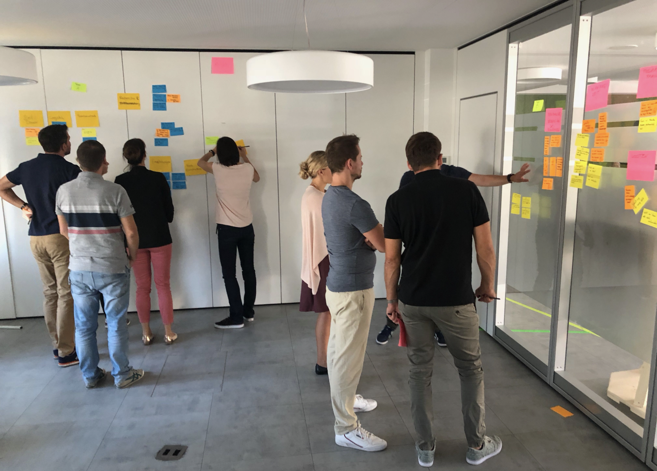 Group of eight people collaborating on a project with colorful sticky notes on whiteboard and glass wall in a modern office.
