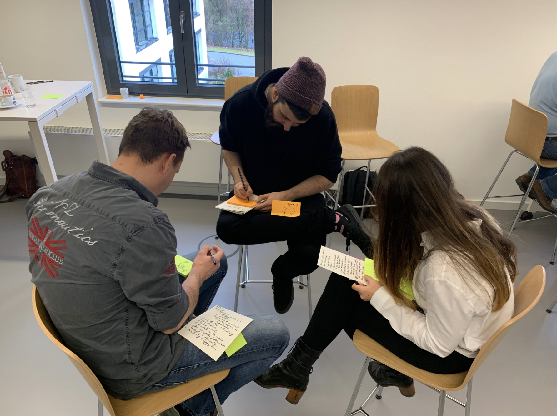Three people sitting in a circle in a classroom, engaging in a discussion and writing on sticky notes and notecards.