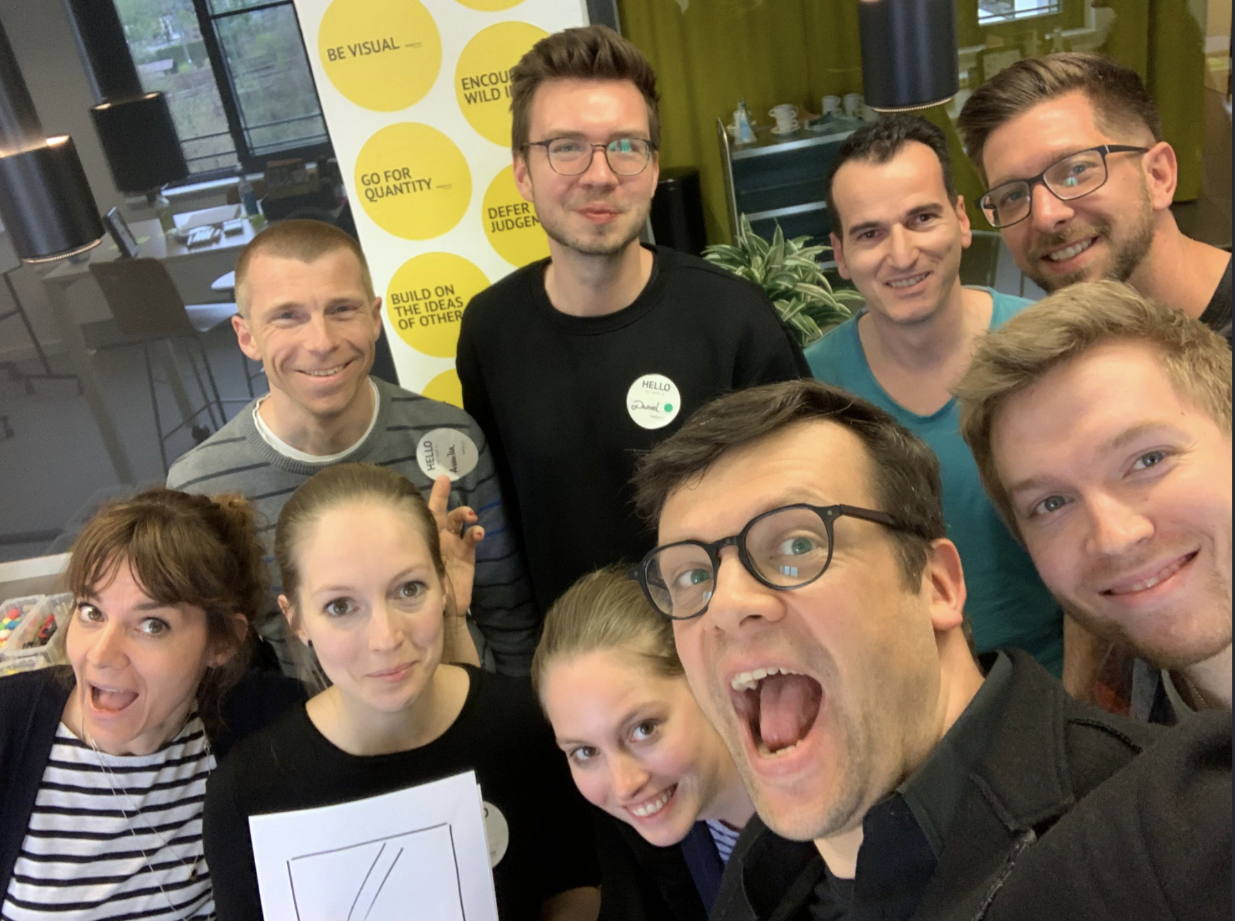 Group of nine smiling people taking a selfie in an indoor setting with a yellow poster in the background displaying motivational phrases.