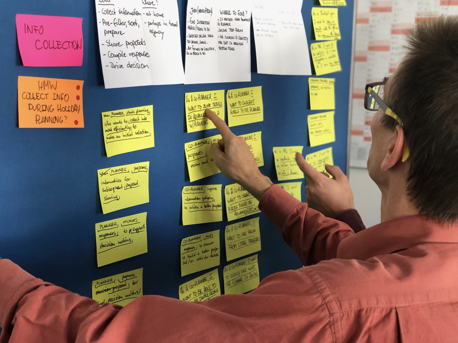 A man with glasses and a red shirt points to yellow sticky notes on a blue board, discussing details about a planning or brainstorming session. There are also white and pink notes on the board.