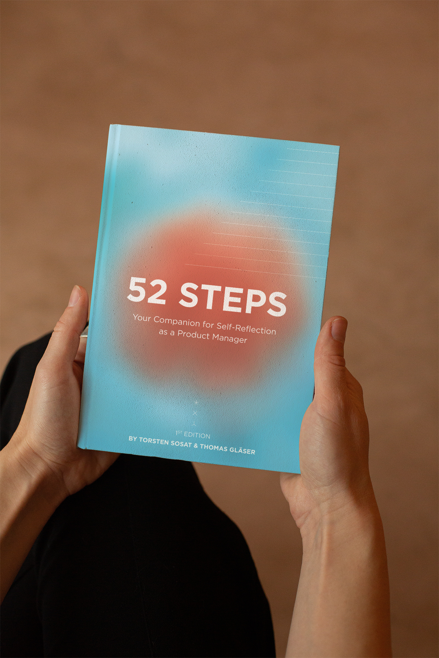A person holding a book titled '52 STEPS: Your Companion for Self-Reflection as a Product Manager,' by Torsten Sosat and Thomas Gläser, with a colorful gradient cover